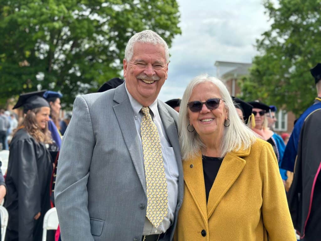 Chp Phillips in grey jacket, light shirt and light gold tie, with Martha Phillips in sunglasses, gold jacket, and dark shirt, in front of campus trees and graduates in gowns