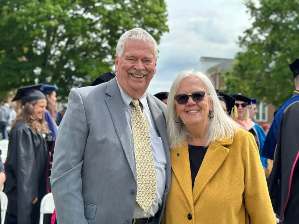Chp Phillips in grey jacket, light shirt and light gold tie, with Martha Phillips in sunglasses, gold jacket, and dark shirt, in front of campus trees and graduates in gowns