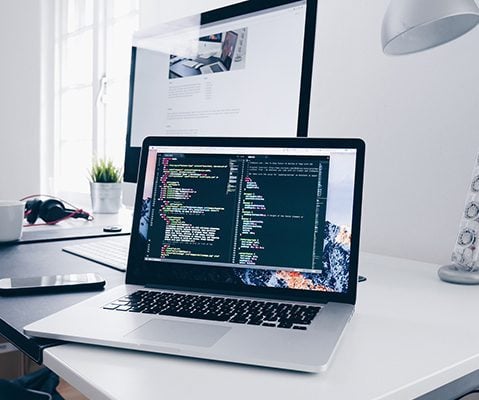 laptop open with coding on screen laying in front of a monitor on a white table