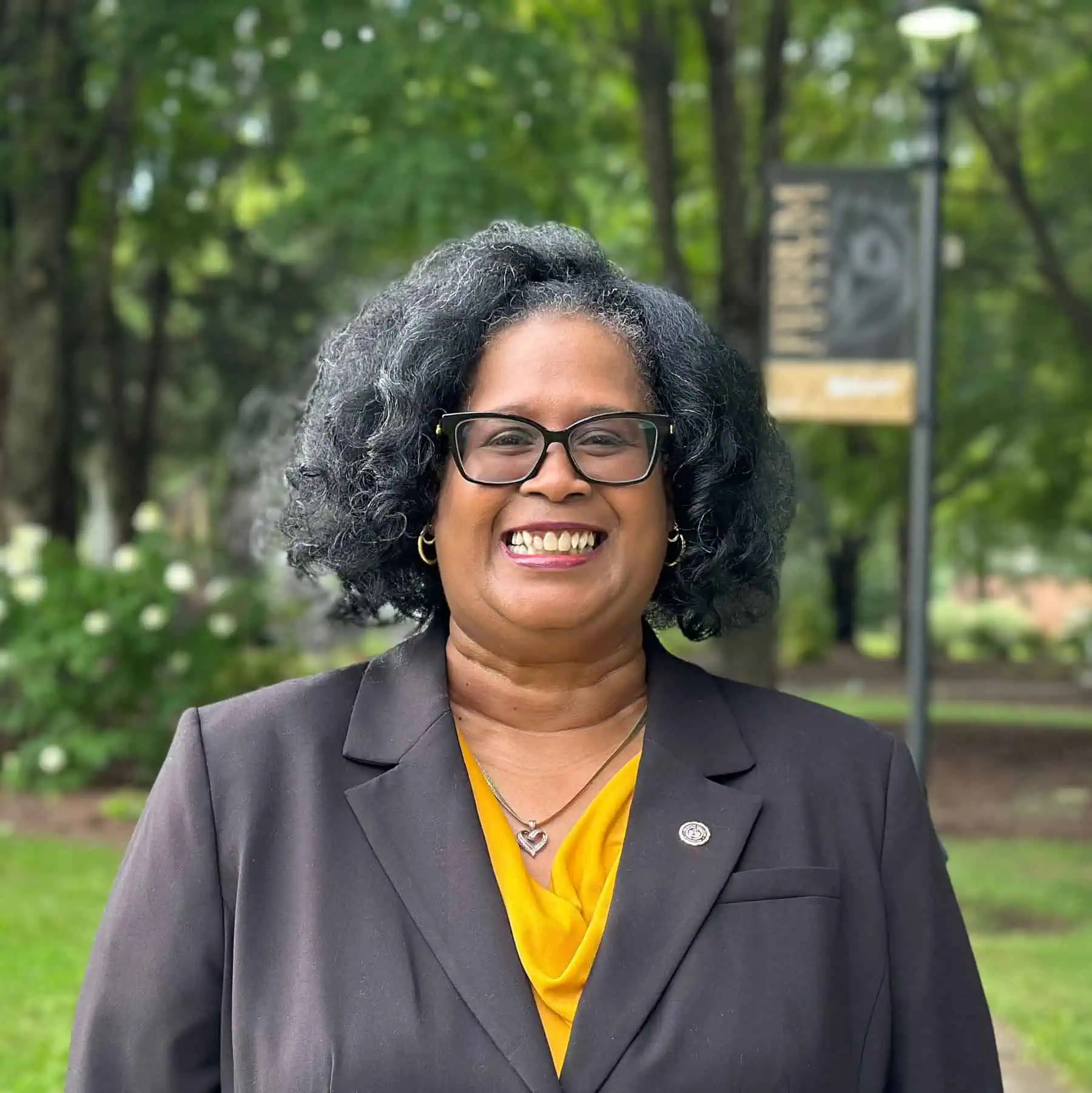 Ferrum College Director of Alumni and Family Relations Corretta Patterson is standing outside on campus wearing glasses and a black blazer over a gold blouse