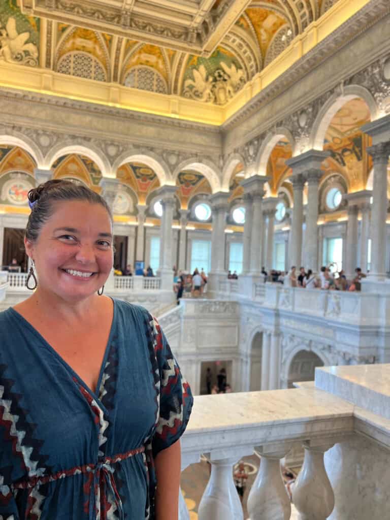 A woman in a blue patterned dress smiles in a grand marble hall with ornate ceilings and columns, filled with visitors.