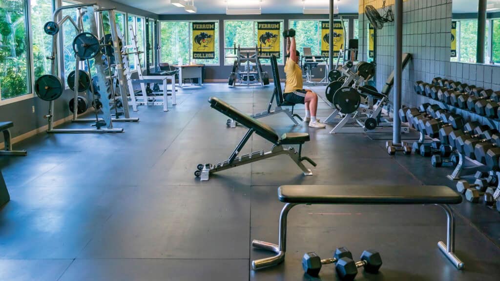 Fitness Center indoors with weight training equipment, shiny floor, big windows, and one person lifting a weight.