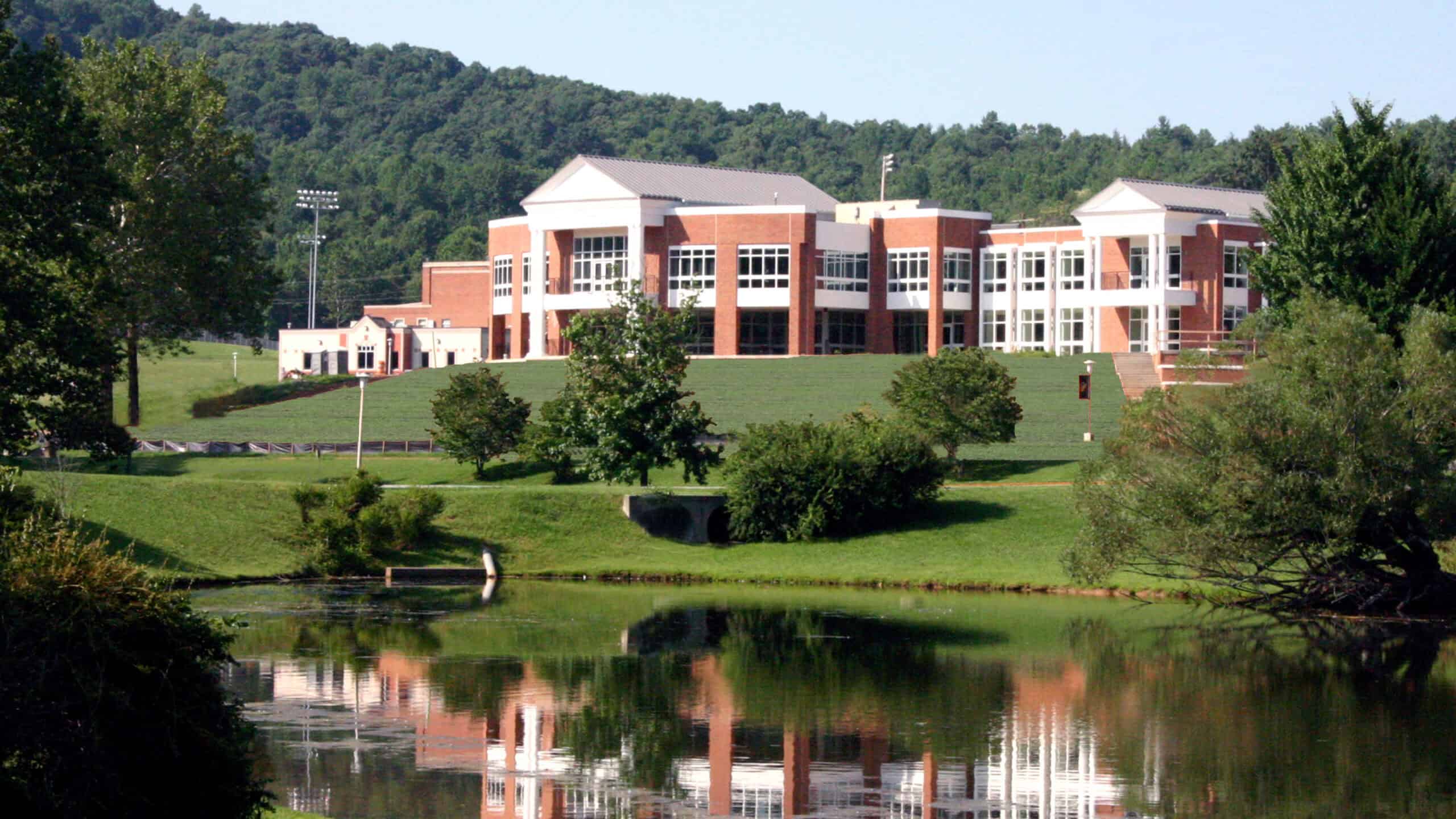 Photo of a red brick with white trim building reflected in a lake with green trees and blue sky