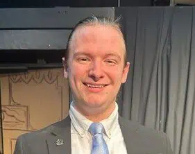 Gage Shelton head shot with hair pulled back, white shirt, grey jacket, and light blue tie, with theatre curtain and door in background