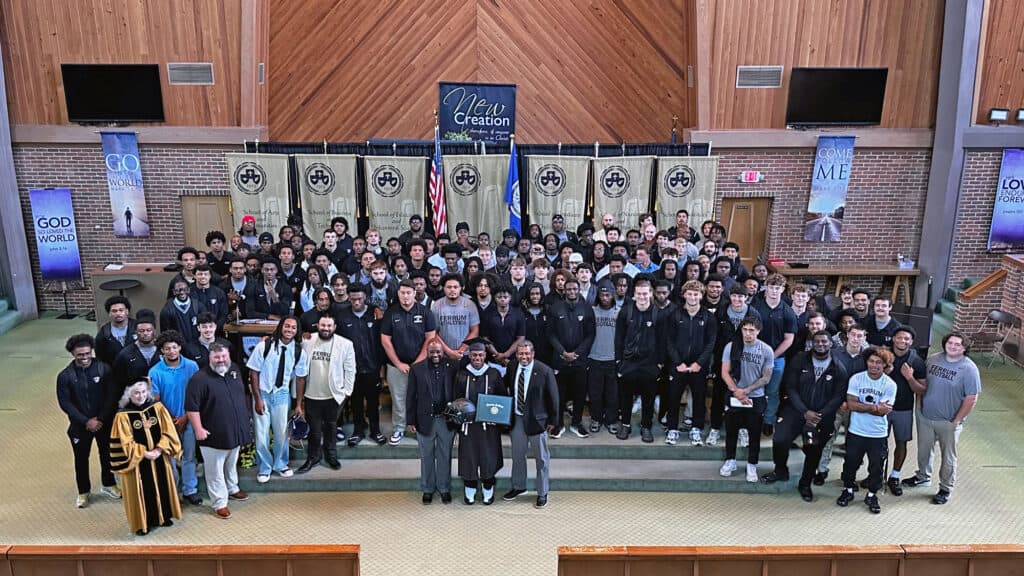 A large group of students, coaches, and faculty poses for a photo in a chapel, with one student holding a diploma and a football helmet in the center foreground.