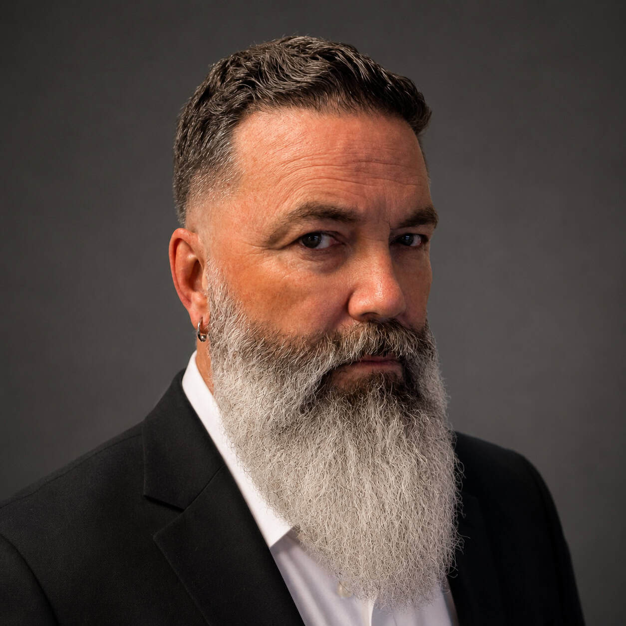Photo of a man with dark hair and a long salt and pepper beard wearing a white dress shirt and a dark suit jacket