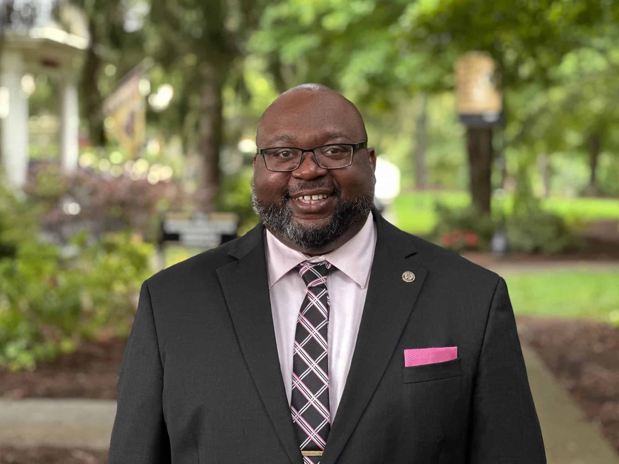 Photograph of Justin Muse wearing a dark suit, pink shirt, plaind tie, and pink pocket square taken outside at Ferrum College on a pretty summer day