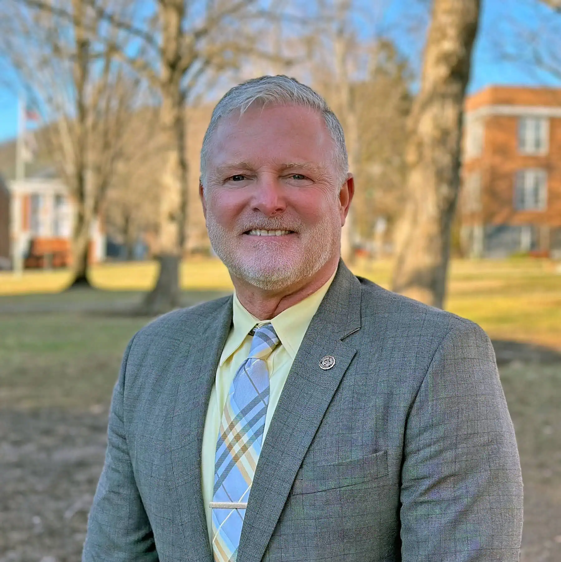 Karl Hatton headshot with man in grey suit and striped tie standing outside in front of brick college buildings and bare trees