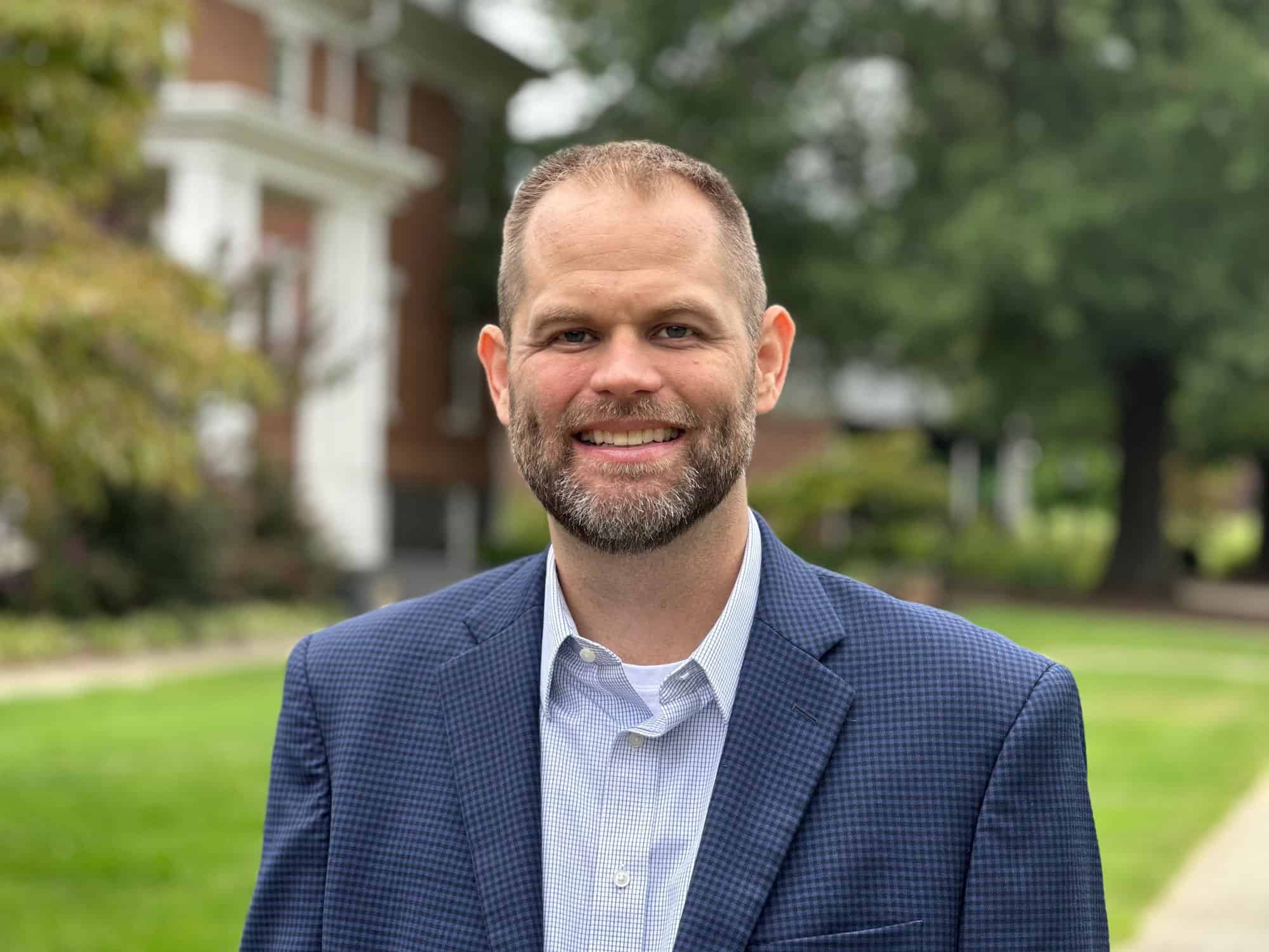 Head shot photo of Kevin Board wearing a blue shiort and blue blazer taken outside on the Ferrum College campus on a pretty summer day with a brick building and green grass and trees in the background