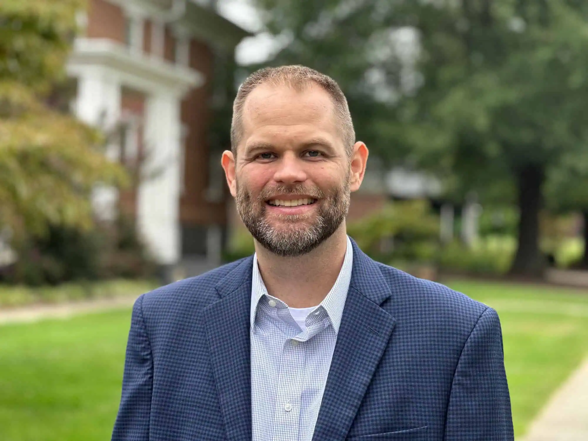 Head shot photo of Kevin Board wearing a blue shiort and blue blazer taken outside on the Ferrum College campus on a pretty summer day with a brick building and green grass and trees in the background