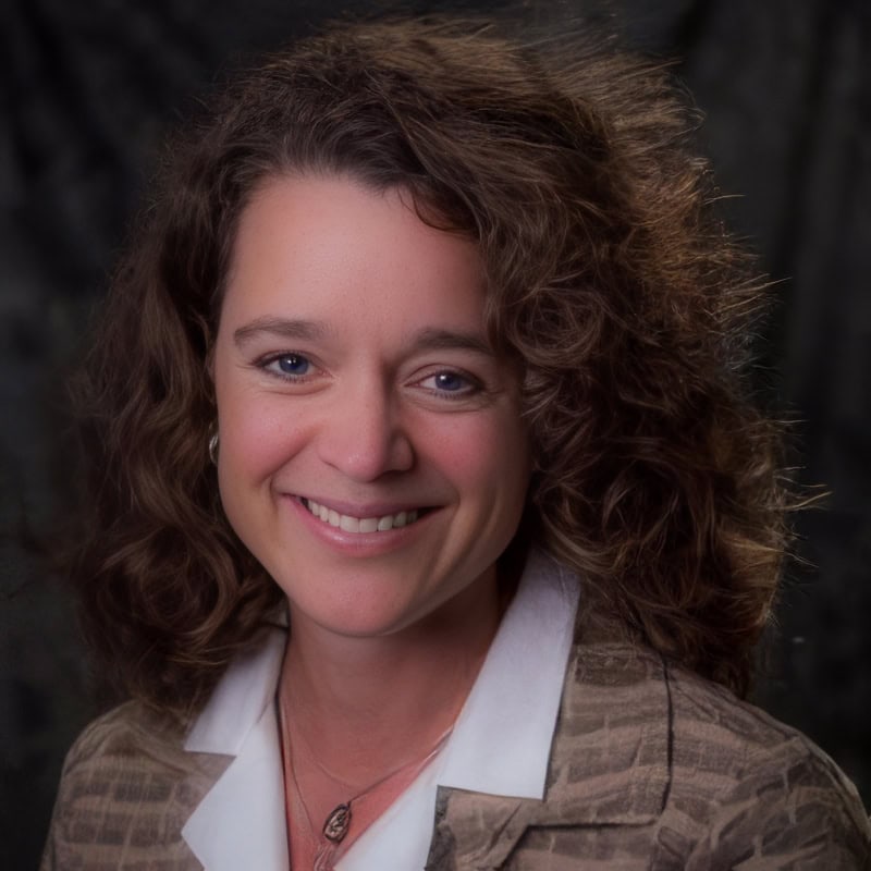 Photo of a woman with long, curly hair, who is wearing a brown textured blazer over a white blouse