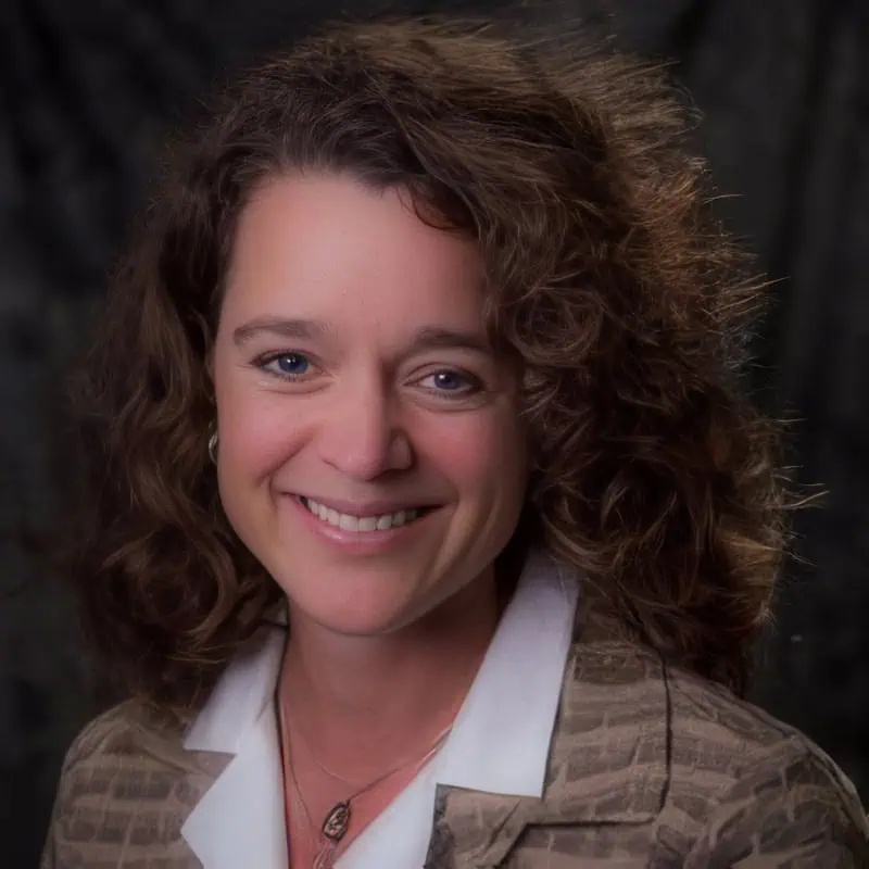 Photo of a woman with long, curly hair, who is wearing a brown textured blazer over a white blouse