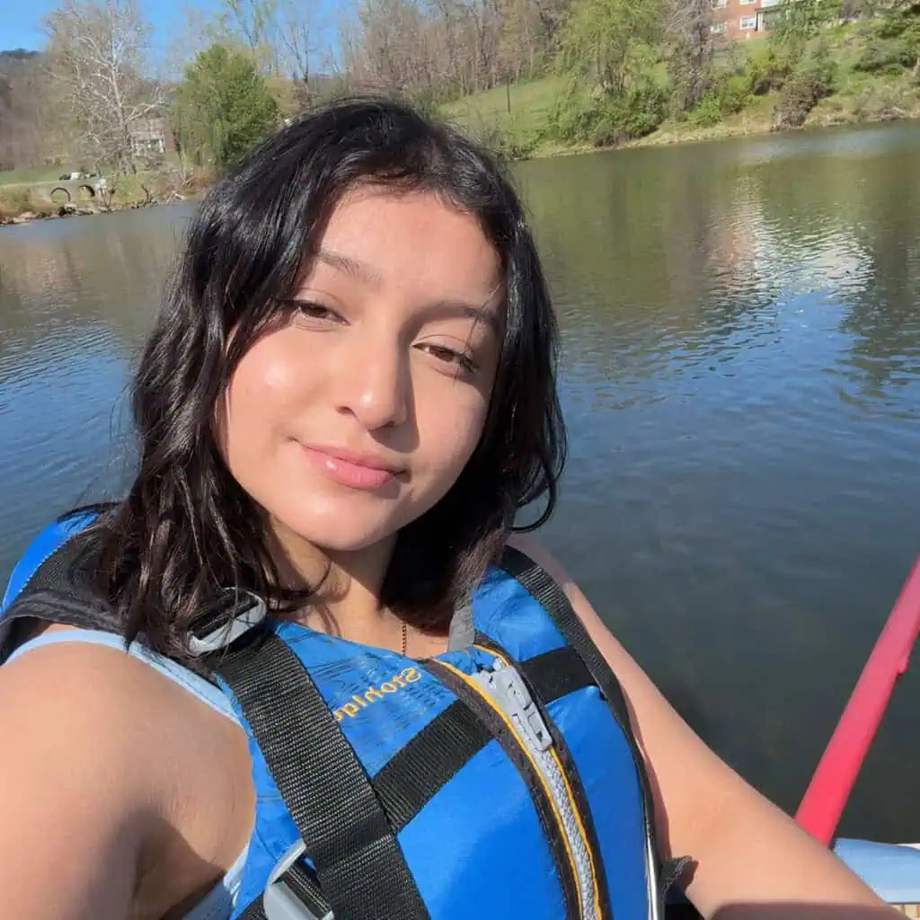 A young woman with black hair wearing a blue life preserver and holding a red paddle is floating on a lake with trees and brick buildings behind her