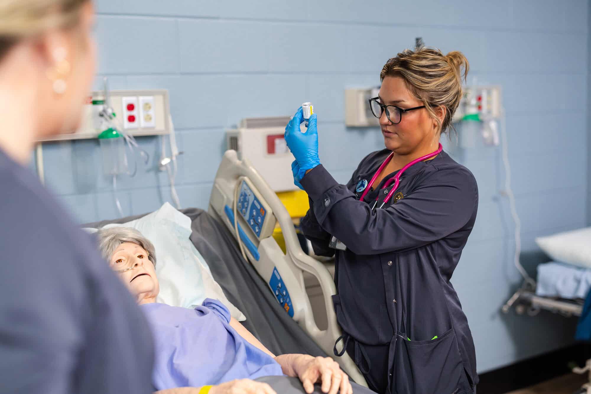 A nurse with glasses, upswept hair, dark shirt, instrument around her neck, and blue gloves holds up a small container. Technical equipment is in front of her on left and behind her.
