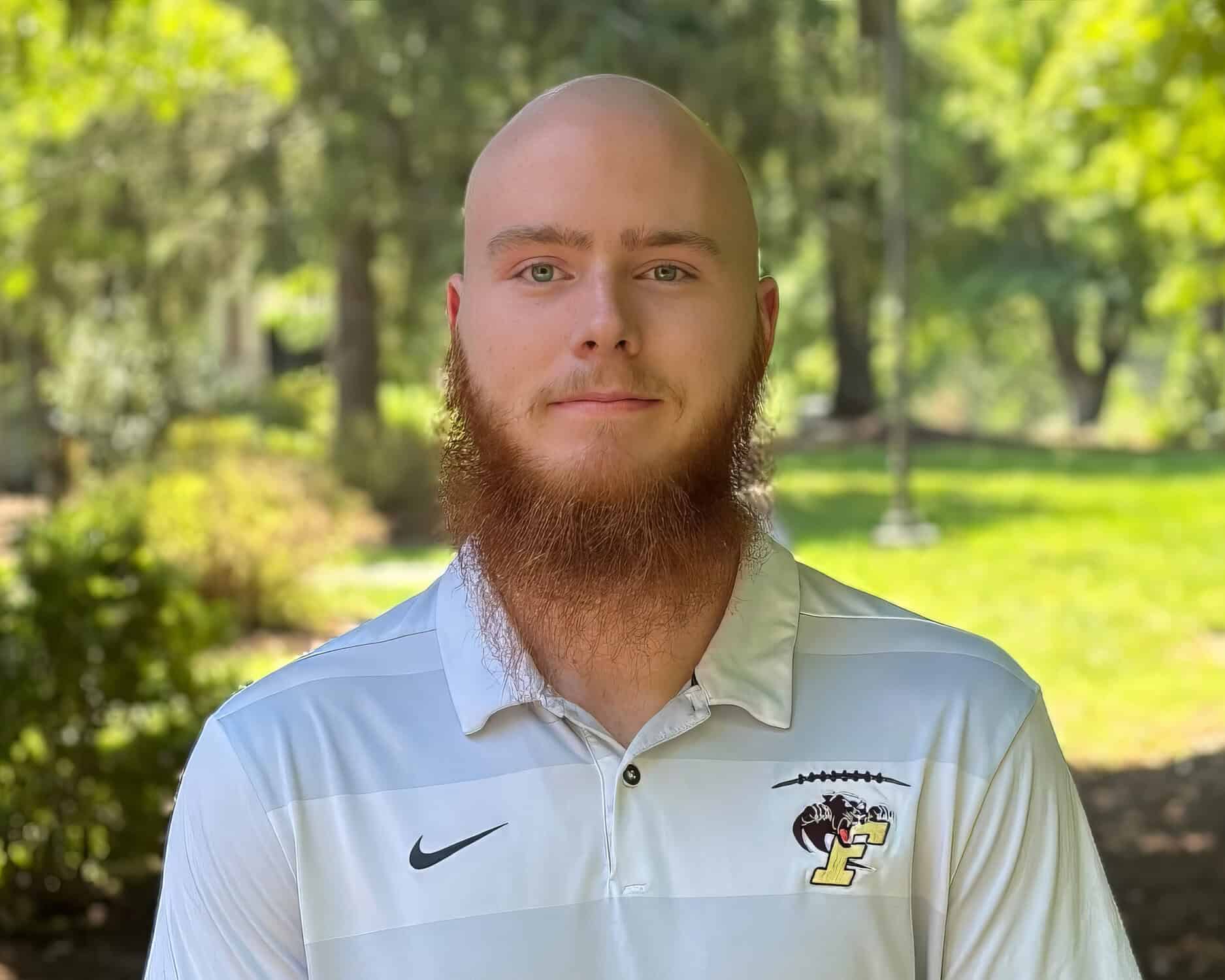 Orion Harris standing in the shade on a sunny day. Trees in the background. He's wearing a white and grey shirt.