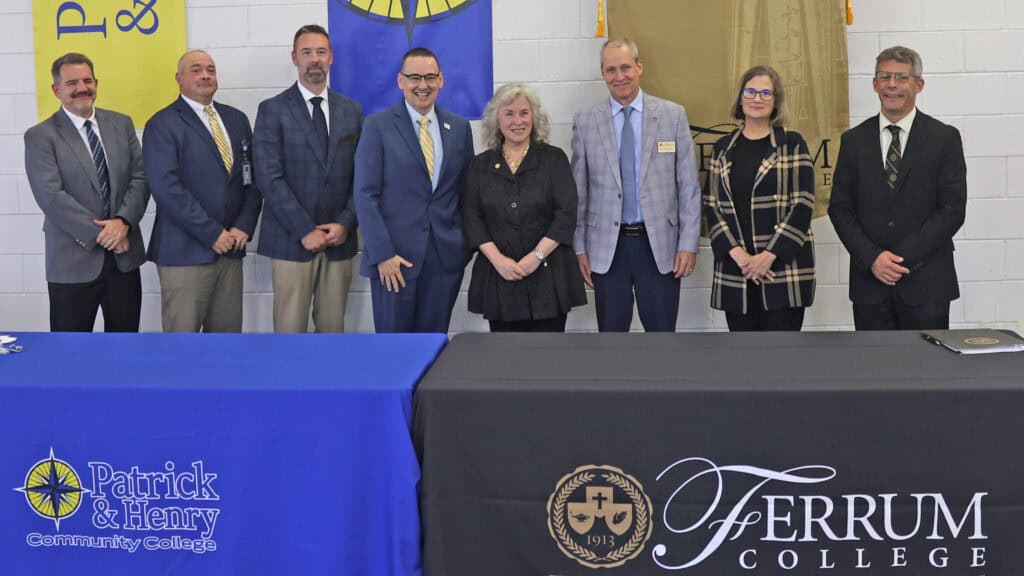 Seven professionals stand in a row behind two tables with college branding, posing for a formal group portrait against a white brick wall background.