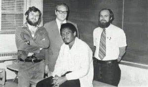 1978 black and white yearbook photo of 4 social science faulty standing in a classroom: Andrew Baskins sitting in front of Richard Smith, Sam Payne, and Ira Colby