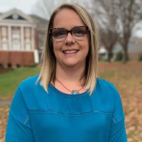 Headshot of Ashley Williams in blue shirt and glasses in front of Sale Theatre.