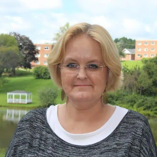 Headshot of Betsy Smith in gray sweater and glasses in front of Adams lake.
