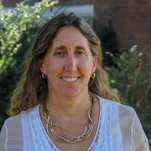 Headshot of Delia Heck in white top with a necklace and earrings in front of bush on campus.