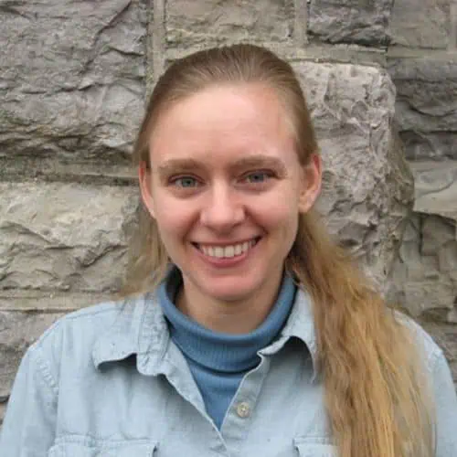 Headshot of Laura Grochowski in blue turtle neck and denim shirt in front of stone wall.