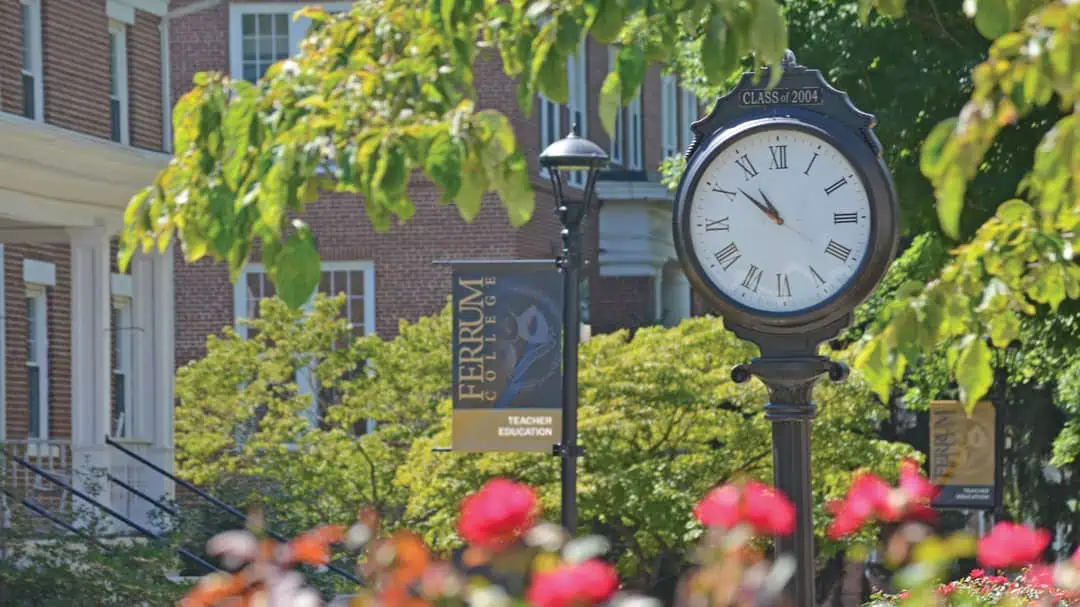Ferrum College campus photo showing an outdoor clock, a teacher education banner, brick buildings, red flowers, and trees