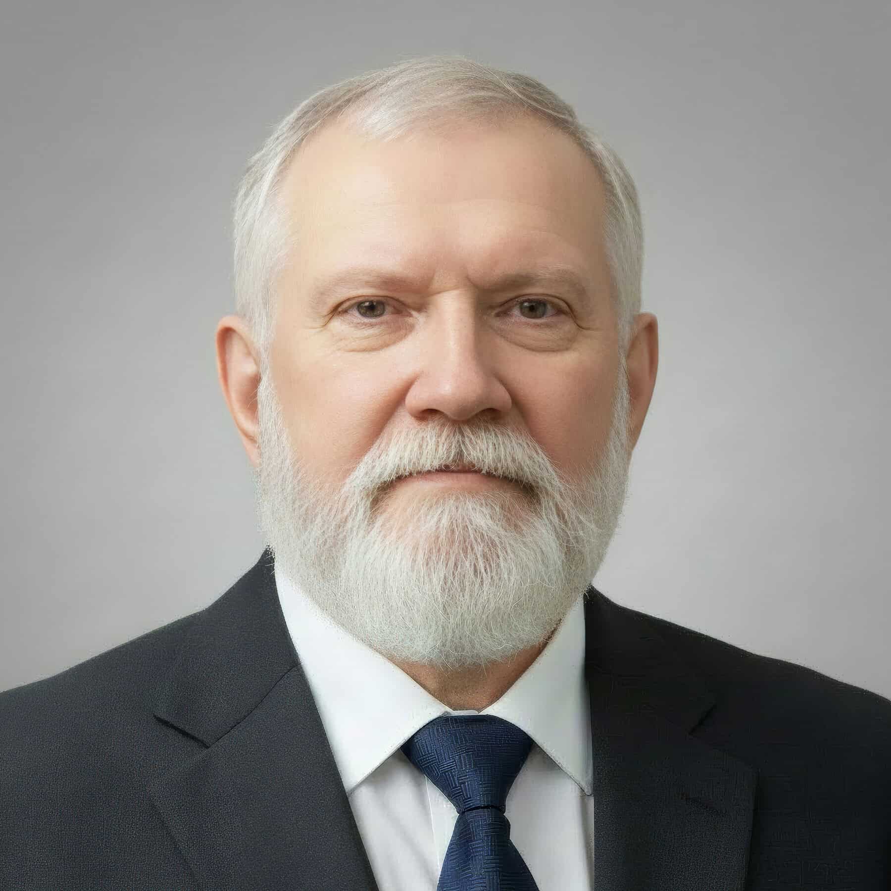 Photo of a white man with gray hair and beard wearing a dark suit, white shirt, and dark blue tie, with a gray background