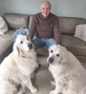 Dr. Ira Colby in jeans and brown shirt on an l-shaped brown sofa with two large white dogs in front