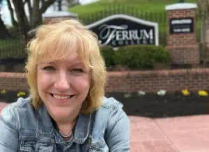 Local author Jane Fenton with blonde hair and blue jacket in front of black and brick Ferrum College entrance sign