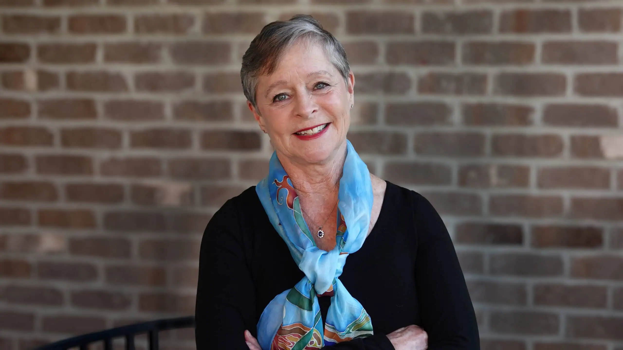 Photo of Janine Latus with arms crossed wearing a black sweater, with a turquoise scarf around her neck, and a brick wall behind her