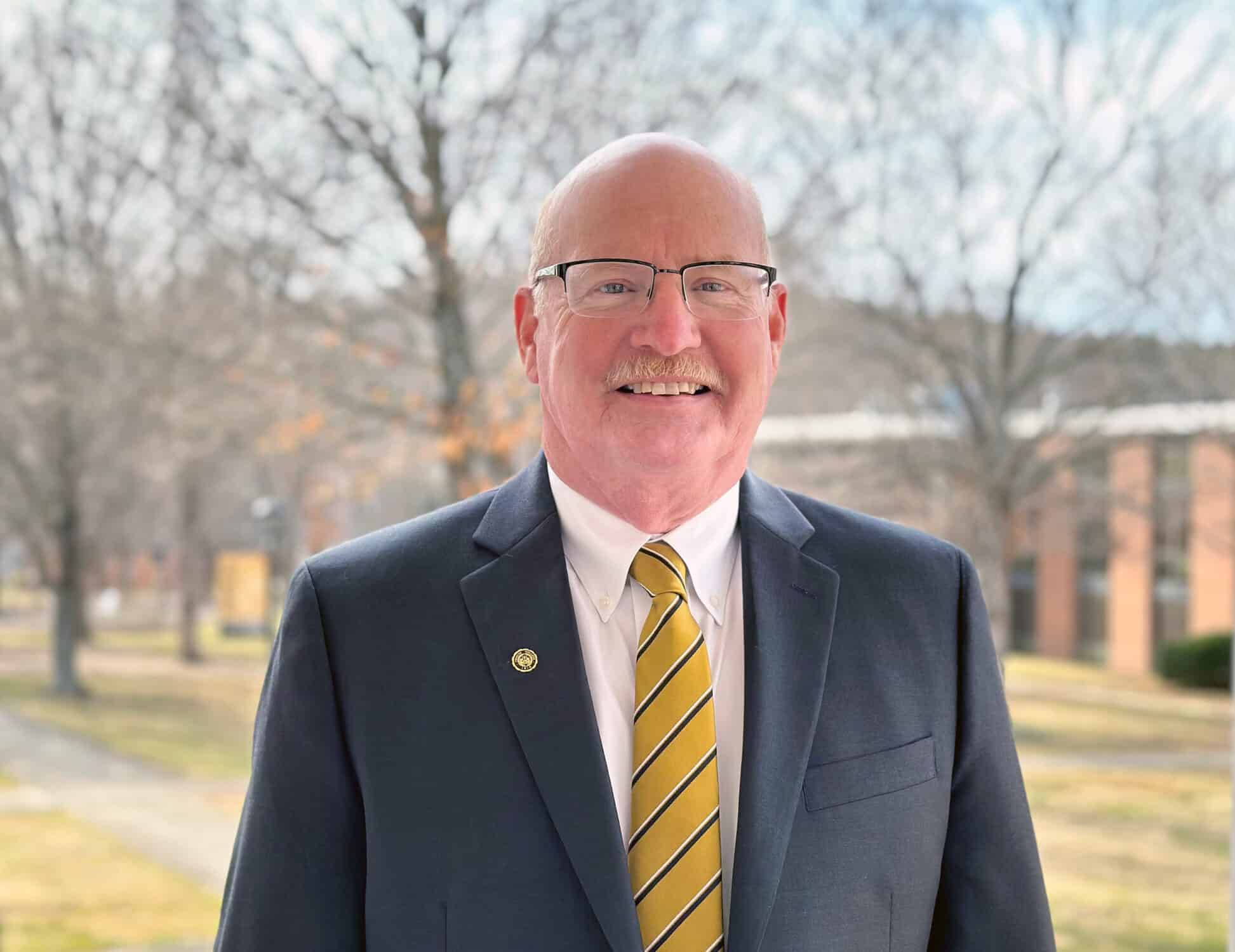 Headshot of smiling man with glasses in dark suit, multicolored tie, and white shirt, with brick building and bare trees in background