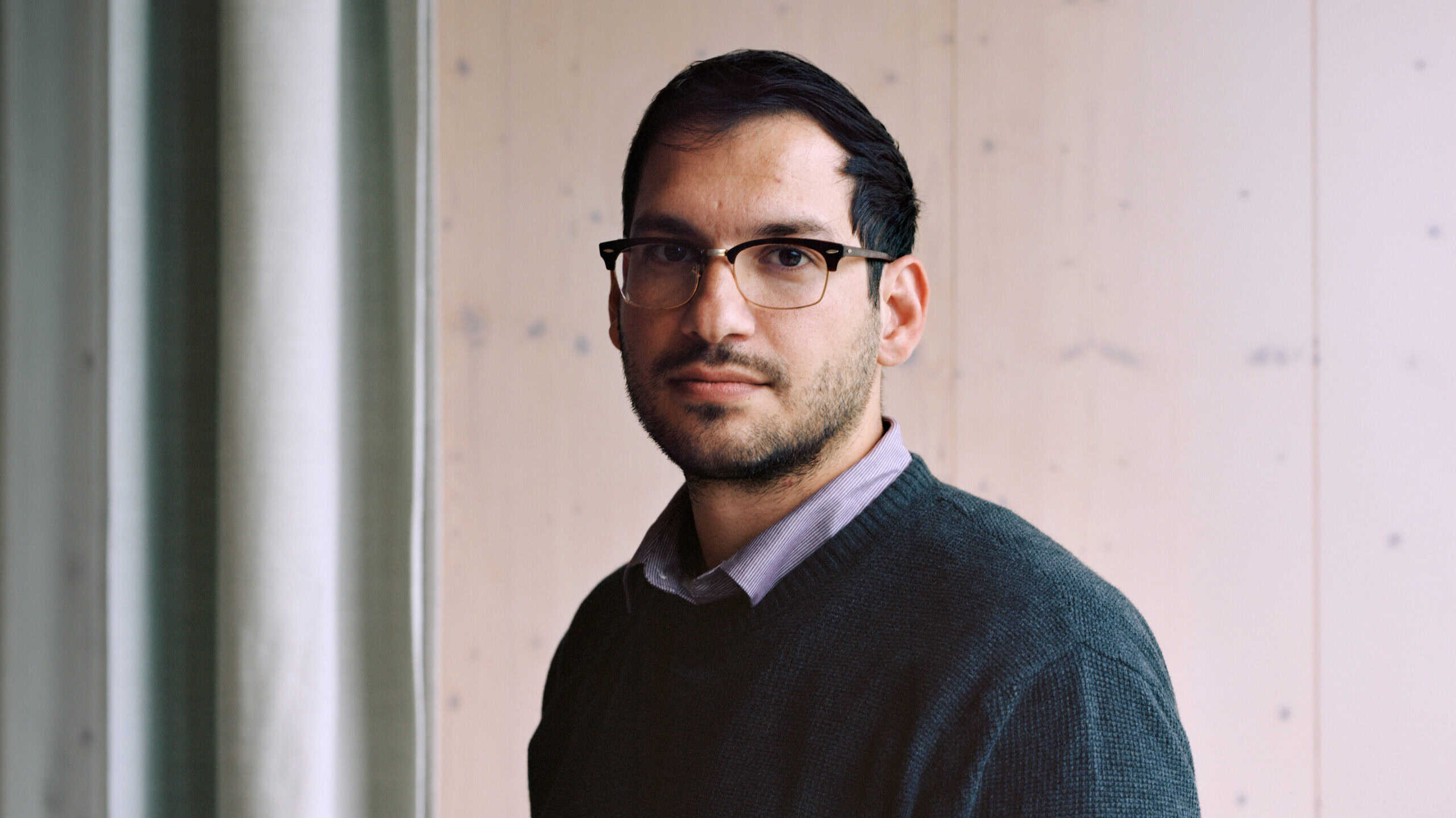 Photo of a dark haired man with a light beard wearing glasses and a navy sweater over a button down shirt