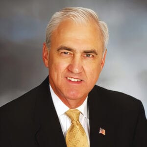 Photo of Jud Mason wearing a black suit, white shirt, gold tie, and US flag lapel pin