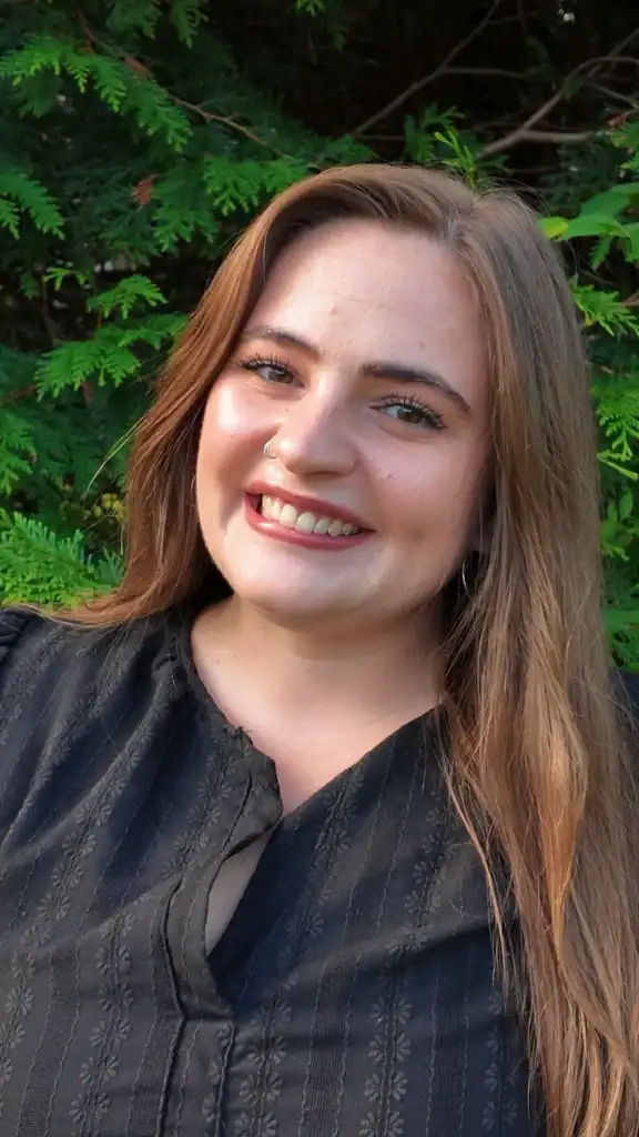 Head shot of recent college graduate in dark blouse, long brown hair, with some foliage behind her