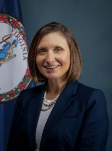 A smiling woman wearing a blue blazer over a white blouse and two necklaces with the Commonwealth of Virginia flag behind her