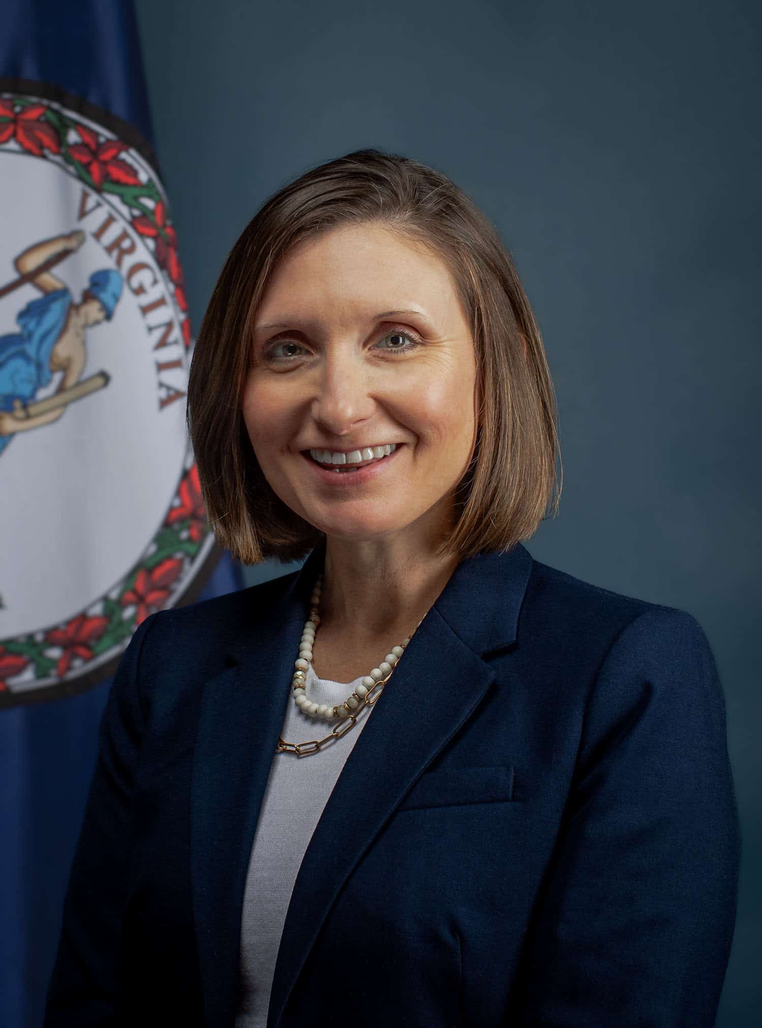 Virginia Secretary of Agriculture Katie Frazier, head shot with part of a state seal behind her. She has chin-length hair, a dark jacket, and white necklace and blouse.