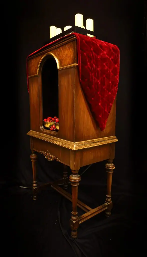 applied gold leaf accents to a large wooden cabinet that holds golden bowls full of cherries with candles on the top and showcases a pitcure